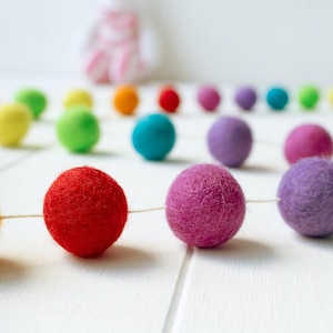 May include: A garland featuring a series of spherical felt balls in various colors including orange, red, purple, yellow, green, and blue, strung together on a thin white cord. The felt balls have a textured, fuzzy surface appearance.
