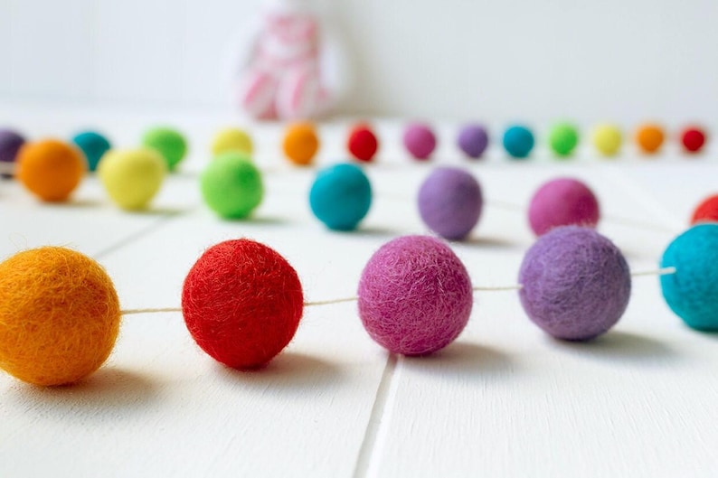 May include: A garland featuring a series of spherical felt balls in various colors including orange, red, purple, yellow, green, and blue, strung together on a thin white cord. The felt balls have a textured, fuzzy surface appearance.