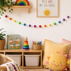 May include: A felt ball garland with rainbow colors, a wooden stacking ring toy, a book titled The Happy Day, a woven basket with crayons, a yellow pom-pom pillow with a sun face, and a framed print with the text You Are My Sunshine.