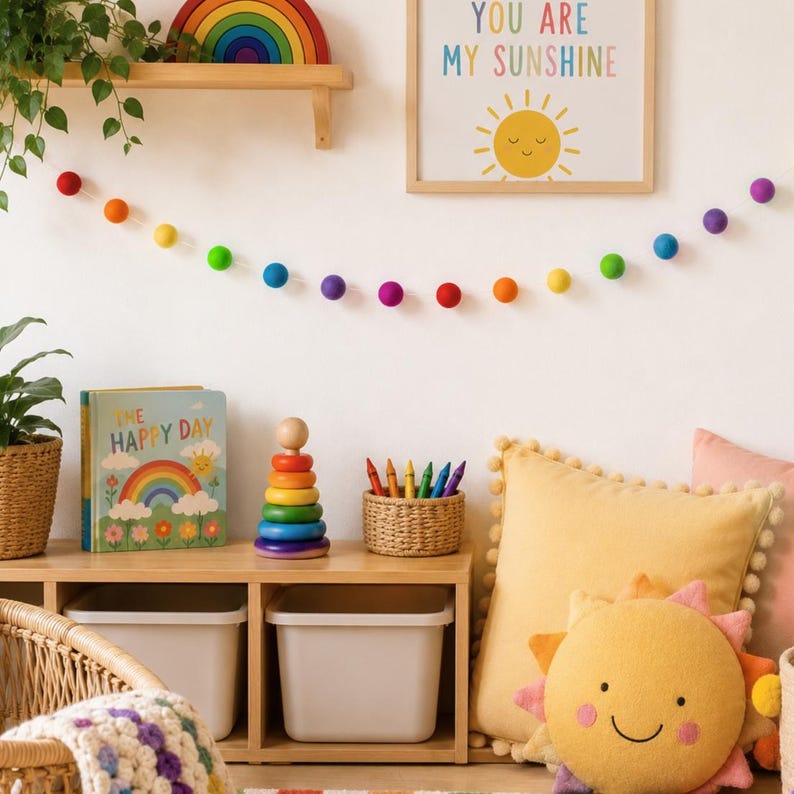 May include: A felt ball garland with rainbow colors, a wooden stacking ring toy, a book titled The Happy Day, a woven basket with crayons, a yellow pom-pom pillow with a sun face, and a framed print with the text You Are My Sunshine.