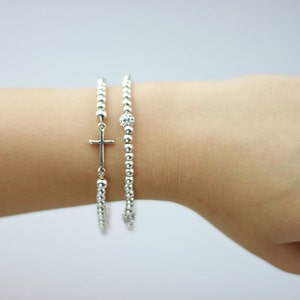 May include: Two silver beaded bracelets on a wrist. One bracelet has a small silver cross charm.