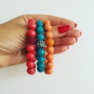 May include: Four colourful beaded bracelets stacked on a hand. The bracelets are red, turquoise, orange, and a darker red. The bracelets have a sparkly bead in the centre.