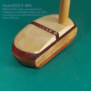 May include: A wooden golf putter with a maple and padauk wood design. The putter has a contrasting red stripe and brass accents. The putter is built on a beefy oak core, sandwiched between two layers of padauk.