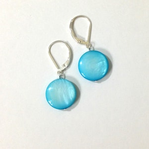 May include: A pair of drop earrings featuring round, bright blue iridescent shell discs suspended from silver-toned metal leverback closures. The smooth, circular shell components display a subtle pearlescent sheen and are attached to the metal findings.