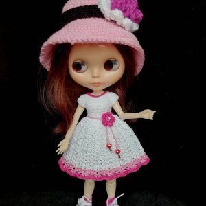 May include: A doll wearing a pink crocheted hat with a white and purple flower, a white crocheted dress with a pink flower and pink trim, and pink and white sneakers.