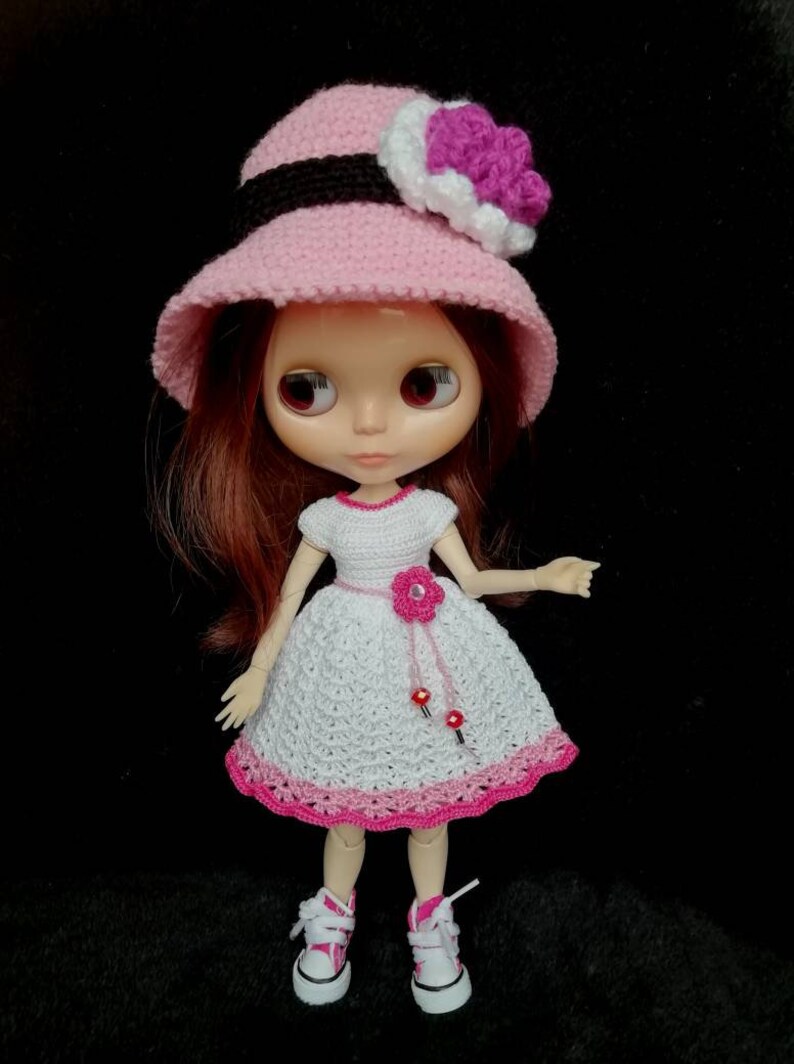 May include: A doll wearing a pink crocheted hat with a white and purple flower, a white crocheted dress with a pink flower and pink trim, and pink and white sneakers.