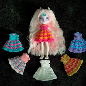 May include: A white doll with pink hair wearing a yellow and pink striped dress. The doll is surrounded by six crocheted dresses in various colours, including pink, blue, white, and purple.