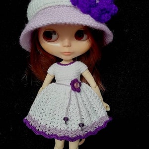 May include: A doll wearing a white crocheted dress with purple trim, a lavender crocheted hat with a purple flower, and white sneakers.