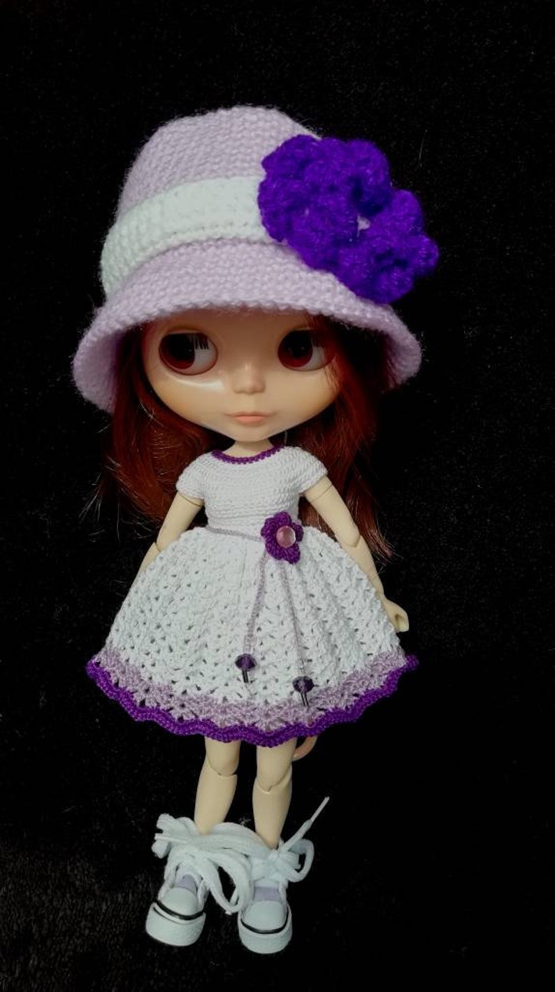 May include: A doll wearing a white crocheted dress with purple trim, a lavender crocheted hat with a purple flower, and white sneakers.