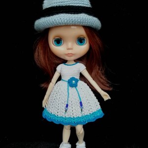 May include: A doll wearing a white crocheted dress with a blue flower and blue beads, a light blue crocheted hat with a black band, and white sneakers.