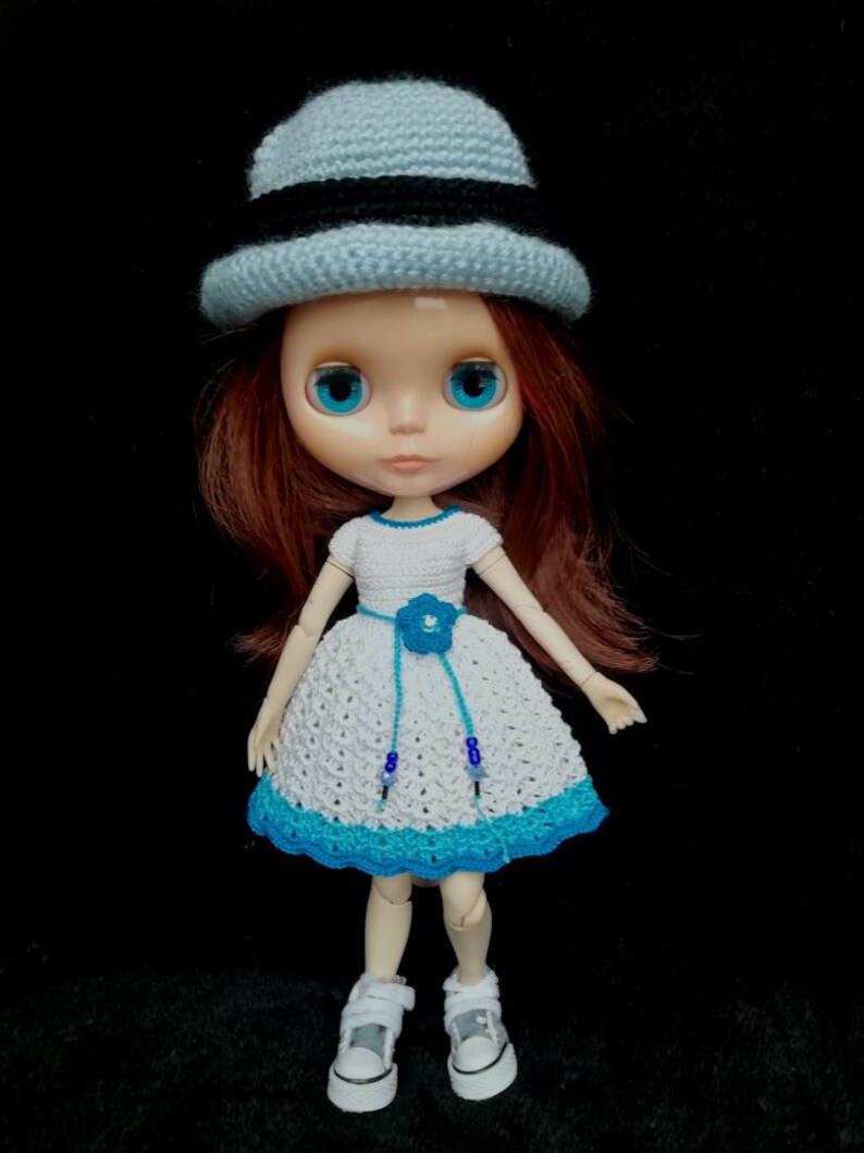 May include: A doll wearing a white crocheted dress with a blue flower and blue beads, a light blue crocheted hat with a black band, and white sneakers.