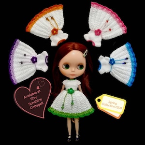 May include: A doll wearing a green and white crocheted dress with a green flower and beads. The doll is surrounded by five crocheted dresses in different colors, including purple, blue, pink, orange, and white. The text "Spring Collection 2020" is on a yellow tag. The text "Available at Etsy Sunshine Cottage8" is on a pink heart.