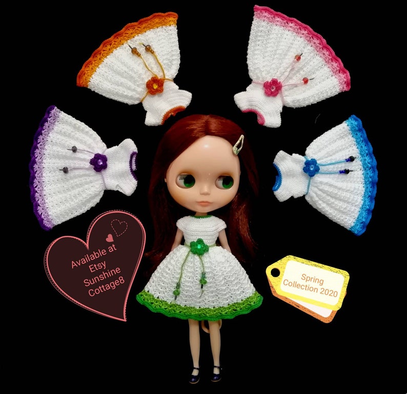 May include: A doll wearing a green and white crocheted dress with a green flower and beads. The doll is surrounded by five crocheted dresses in different colors, including purple, blue, pink, orange, and white. The text "Spring Collection 2020" is on a yellow tag. The text "Available at Etsy Sunshine Cottage8" is on a pink heart.
