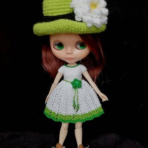 May include: A doll wearing a white crocheted dress with green trim, a green crocheted hat with a white flower, and brown shoes.