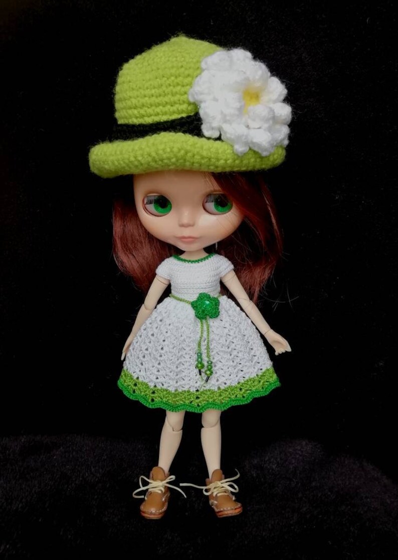 May include: A doll wearing a white crocheted dress with green trim, a green crocheted hat with a white flower, and brown shoes.