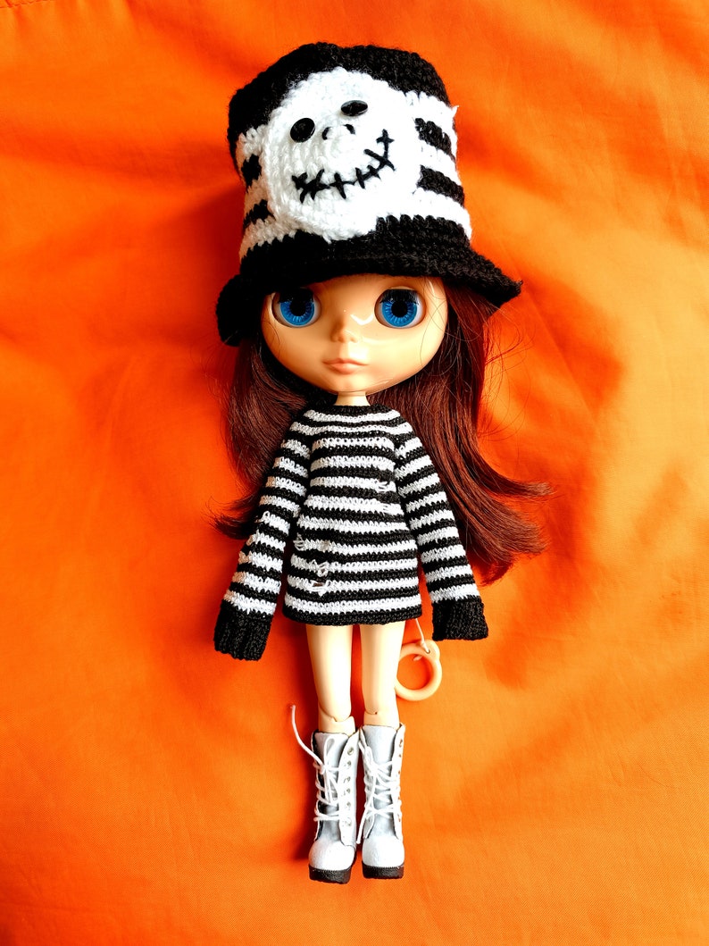 May include: A doll with long brown hair wearing a black and white striped sweater and a black and white skull hat. The doll has blue eyes and is wearing white boots.
