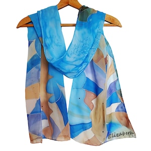 Puede incluir: Bufanda estampada en tonos azules, beige y morados, colgada de una percha de madera. La bufanda presenta un diseño de acuarela. El nombre "Elizabeth" está escrito en la esquina inferior derecha. La bufanda es de un material ligero.