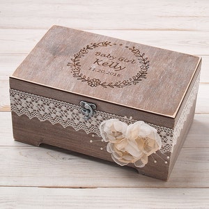May include: A wooden keepsake box with a lace trim and a flower embellishment. The lid is engraved with the text "Baby Girl Kelly 11.20.2016" inside a floral wreath.