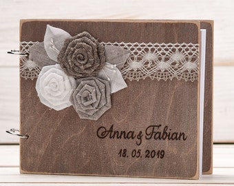 Op de afbeelding: Een houten gastenboek met een bruine houten kaft en een kantversiering. De kaft is versierd met drie stoffen bloemen in grijstinten en wit. Het boek is gepersonaliseerd met de tekst "Anna & Fabian 18.05.2019".