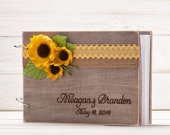 May include: A wooden guest book with a burlap and lace ribbon, decorated with three yellow felt sunflowers. The book is personalized with the names "Meagan & Brandon" and the date "May 18, 2019".