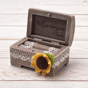 May include: A rustic wooden ring box with a sunflower design. The lid is open and the inside is lined with burlap. The lid is engraved with the words "Will you marry me - Charles".