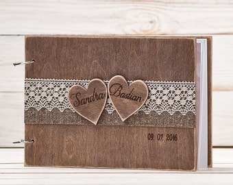 May include: A wooden photo album with a rustic brown finish. The album has a lace trim and two wooden hearts with the names "Sandra" and "Bastian" engraved on them. The date "09.07.2016" is also engraved on the front of the album.