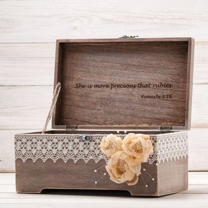 May include: A wooden box with a lace trim and a floral embellishment. The lid is open and the inside is engraved with the text "She is more precious than rubies. Proverbs 3:15."