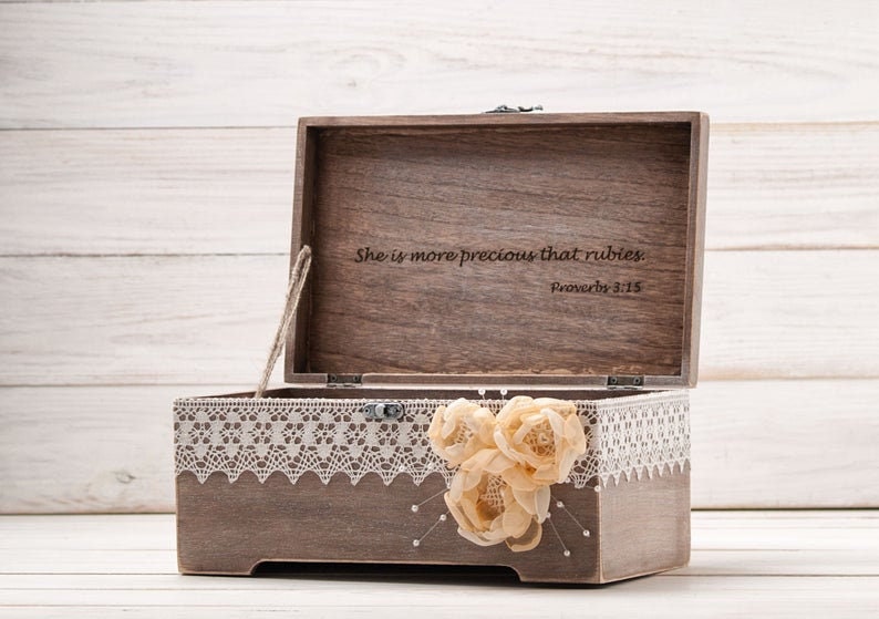 May include: A wooden box with a lace trim and a floral embellishment. The lid is open and the inside is engraved with the text "She is more precious than rubies. Proverbs 3:15."