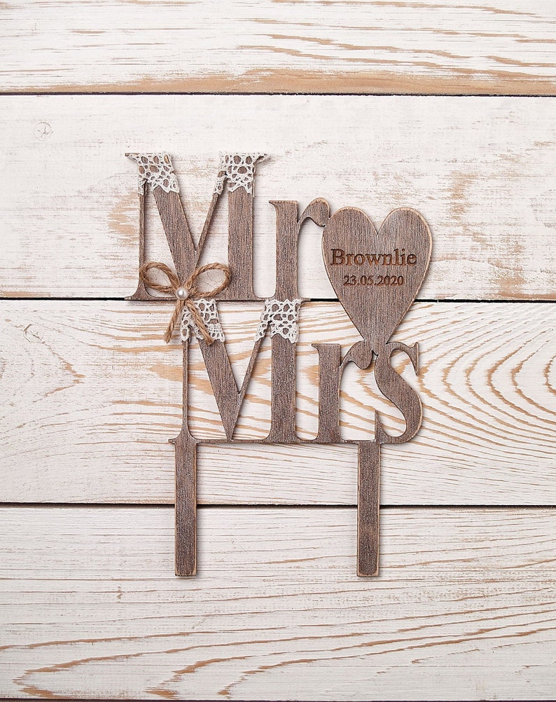 Puede incluir: Topper de pastel de madera "Mr & Mrs" con una etiqueta en forma de coraz&oacute;n que dice "Brownlie 23.05.2020". El topper est&aacute; decorado con encaje y un lazo marr&oacute;n.