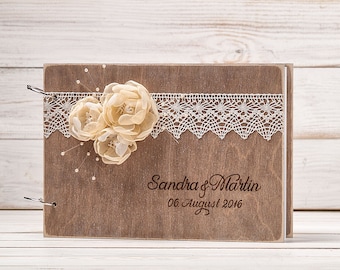 May include: A wooden guest book with a lace trim and three beige fabric flowers. The book is engraved with the text "Sandra & Martin 06 August 2016".