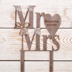Puede incluir: Topper de pastel de madera con las palabras "Mr" y "Mrs" en un estilo r&uacute;stico. El "Mr" tiene un peque&ntilde;o lazo y el "Mrs" tiene un coraz&oacute;n con el texto "Brownlie 23.05.2020" en &eacute;l.