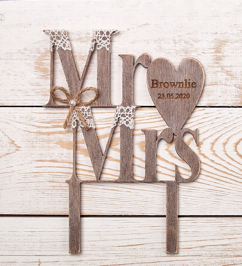 Puede incluir: Topper de pastel de madera con las palabras "Mr" y "Mrs" en un estilo r&uacute;stico. El "Mr" tiene un peque&ntilde;o lazo y el "Mrs" tiene un coraz&oacute;n con el texto "Brownlie 23.05.2020" en &eacute;l.