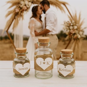 Peut inclure: Trois bouteilles en verre avec des bouchons en liège, remplies de sable et décorées de ficelle et d'étiquettes en forme de cœur. Les noms "Janean" et "Kristian" sont visibles sur deux des étiquettes, et les initiales "J K" sur la troisième. Une cérémonie de mariage est en arrière-plan.