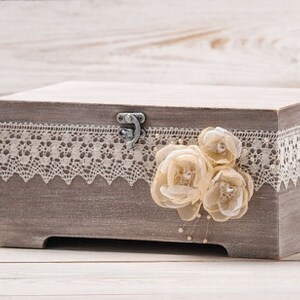 May include: A gray wooden box with a lace trim and a floral embellishment. The box has a hinged lid with a metal clasp.