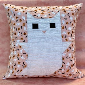 Può includere: Un cuscino quadrato trapuntato con un disegno di gatto. Il volto del gatto è bianco con occhi quadrati neri e un naso rosa. Il bordo e lo sfondo del cuscino presentano un tessuto rosa chiaro con un motivo di fantasmi, pipistrelli e zucche.
