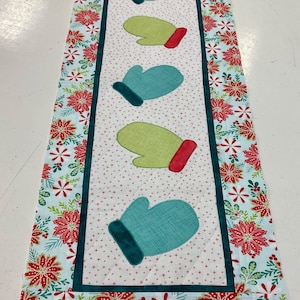 May include: A white table runner with a green and blue mitten design. The runner has a red and white floral border.