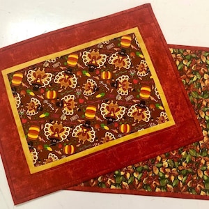 May include: Two rectangular placemats. One has a brown background with turkeys, pumpkins, and text. The other has autumn leaves in brown, green, and orange. Both have a red border. Ideal for a Thanksgiving table setting.
