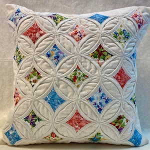 Puede incluir: Cojín decorativo cuadrado con un diseño acolchado. El cojín presenta un fondo gris claro con un patrón de círculos entrelazados. Cada círculo enmarca un parche en forma de diamante de tela floral en rojo, azul y verde.