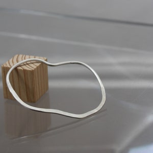 May include: A silver bangle bracelet with an irregular, wavy shape. The bracelet is resting on a wooden block.