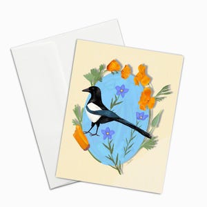 May include: Greeting card featuring a black and white magpie bird illustration. The bird is surrounded by orange flowers, blue flowers, and green foliage against a light blue background. Includes a white envelope.