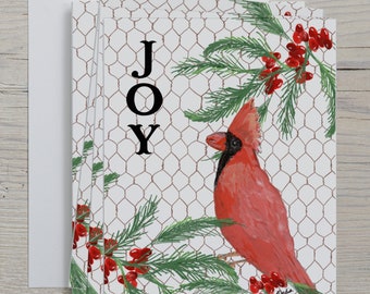 May include: A stack of greeting cards featuring a watercolor painting of a red cardinal bird perched on a branch with red berries and green needles. The word "JOY" is printed in black. The background has a chicken wire pattern.