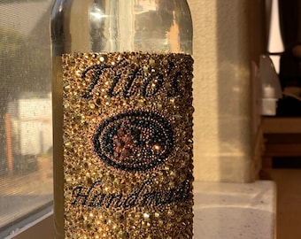 Può includere: Una bottiglia di vetro trasparente con un'etichetta dorata e nera ricoperta di strass. L'etichetta riporta la scritta "Tito's Handmade Vodka".