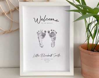 May include: A white framed print with the text "Welcome to the World" and a pair of baby footprints in black ink. The print also includes the name "Lottie Bluebell Smith", the date "1st January 2021", the weight "6lb 13oz", and the time "12:53".