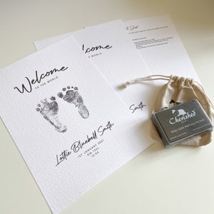 May include: A white card with black text that reads "Welcome to the World" and features two black baby footprints. The card also includes the name "Lottie Bluebell Smith", the date "1st January 2021", the weight "6lb 13oz", and the time "12:53". A second white card with black text reads "Welcome to the World". A small black ink pad with the text "Cherished Baby Baby Safe Memory Ink Pad" is in a small burlap bag.