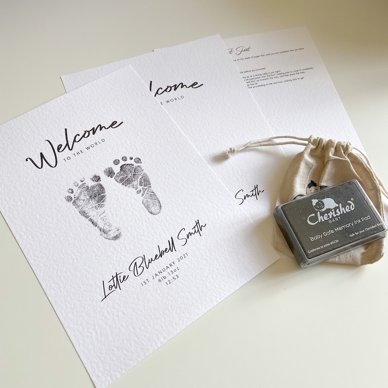 May include: A white card with black text that reads "Welcome to the World" and features two black baby footprints. The card also includes the name "Lottie Bluebell Smith", the date "1st January 2021", the weight "6lb 13oz", and the time "12:53". A second white card with black text reads "Welcome to the World". A small black ink pad with the text "Cherished Baby Baby Safe Memory Ink Pad" is in a small burlap bag.