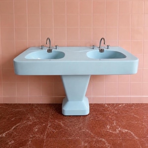 May include: A light blue double pedestal sink featuring two integrated basins, each equipped with a chrome tap and two matching handles. The sink is mounted against a wall covered in square peach-coloured tiles, positioned above a reddish-brown floor.