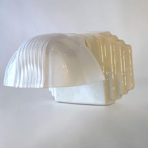 Może przedstawiać: Błyszczący biały kinkiet ceramiczny ze stopniowaną, prostokątną podstawą i zakrzywionym, żebrowanym kloszem wystającym na zewnątrz. Powierzchnia ma gładkie, odblaskowe wykończenie z subtelnymi różnicami odcieni na całej geometrycznej formie.