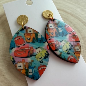 May include: A pair of teardrop-shaped earrings featuring a colorful print of cartoon cars. The earrings have gold-colored circular studs and are displayed on a white card. The design includes various car characters in a vibrant, playful style.