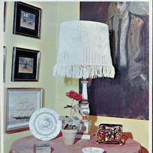 May include: A white macrame lampshade with a fringe trim hangs from a white lamp stand. A red rose in a terracotta pot sits on a table with a small wooden box and a decorative plate.