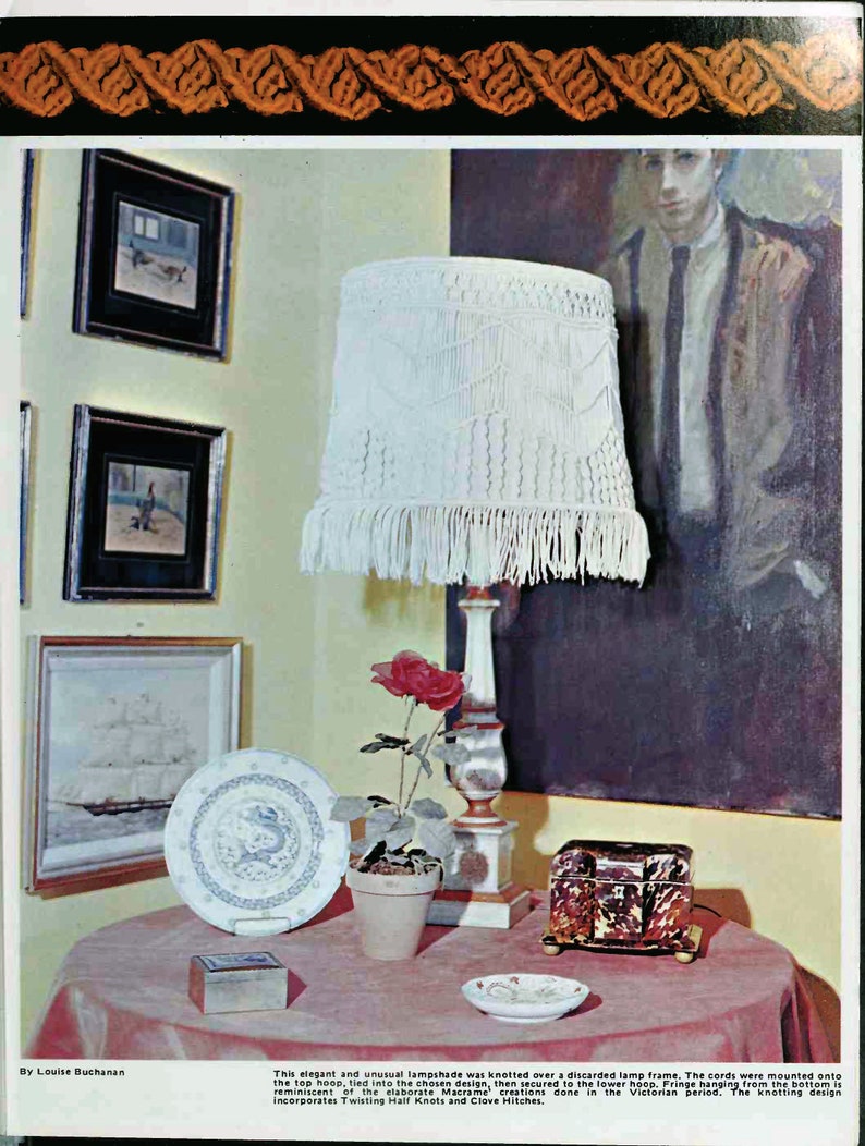 May include: A white macrame lampshade with a fringe trim hangs from a white lamp stand. A red rose in a terracotta pot sits on a table with a small wooden box and a decorative plate.
